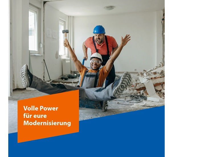 Zwei lachende Männer in Arbeitskleidung auf einer Baustelle im Haus. Der eine Mann schiebt den anderen auf einer Sackkarre.
