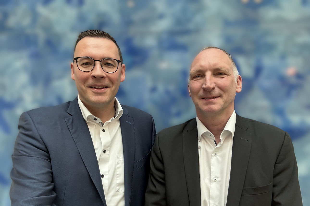 Zwei Individualkundenberater stehen vor einer blauen Wand und lächeln in die Kamera