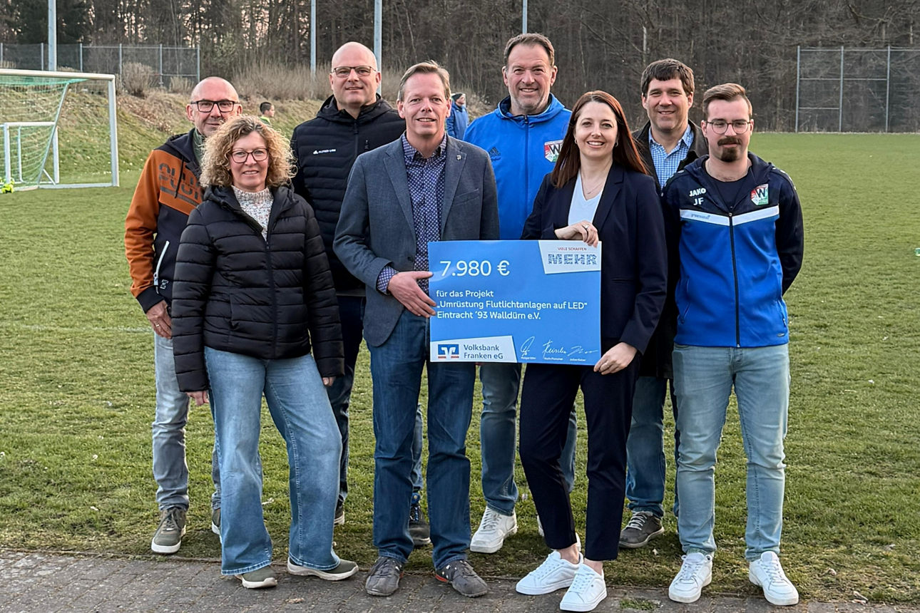 Acht Personen auf einem Fußballplatz; eine Gruppe in Vereinskleidung hält einen großen Spendenscheck.