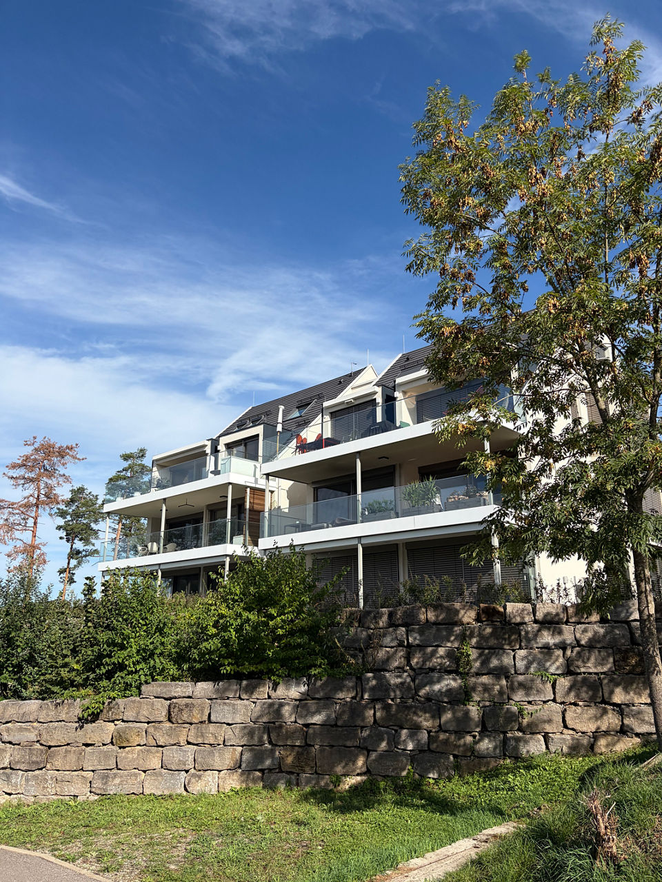 Ansicht eines modernen, hellen Mehrfamilienhauses mit mehreren Balkonen, großen Fenstern und Hecke im Vordergrund. 