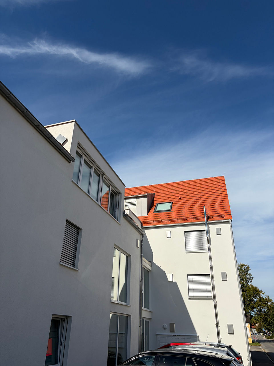 Ansicht eines mehrstöckigen Wohngebäudes mit grauer und weißer Fassade, roten Ziegeldächern, Dachfenstern und geparkten Autos