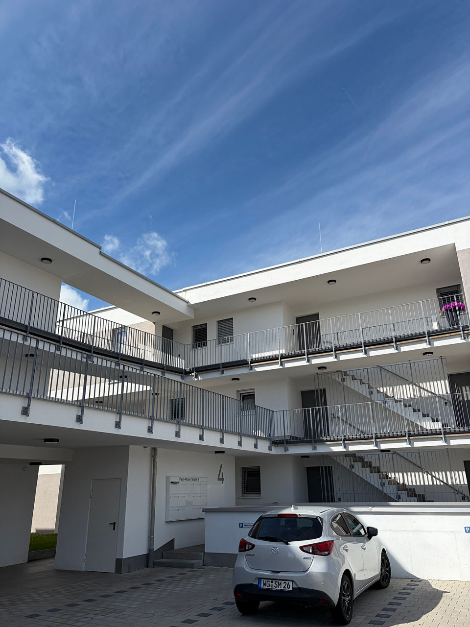 Mehrstöckiges, modernes Wohngebäude mit weißen Fassaden, grauen Metallgeländern, außenliegenden Treppen und Laubengängen.