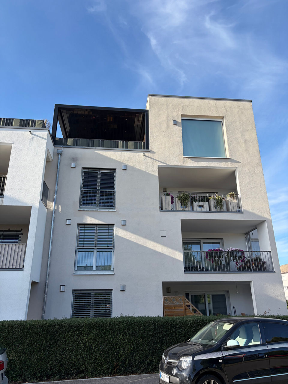 GartenMehrstöckiges Wohnhaus mit Balkonen, weiß-beiger Fassade und geparktem Auto unter blauem Himmel.