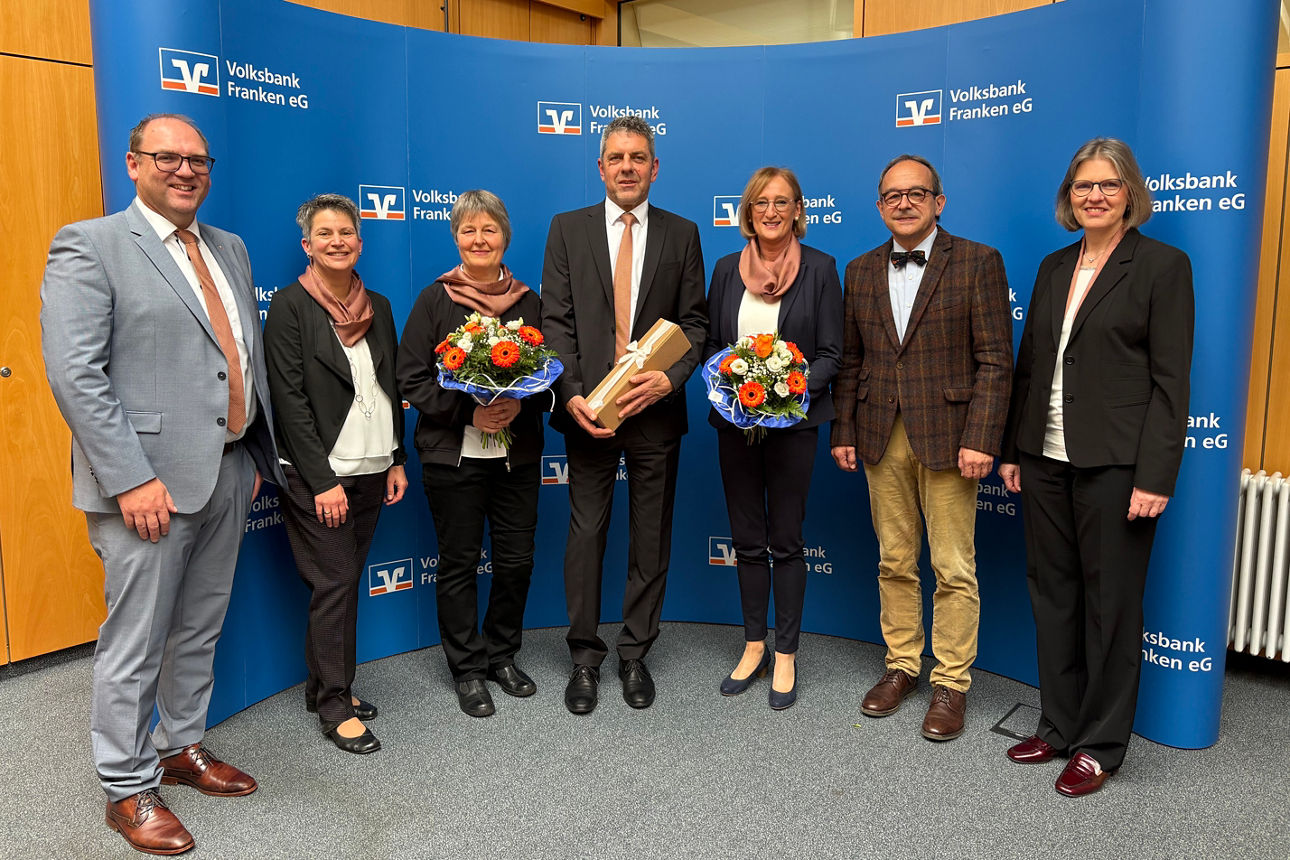 Sieben Personen posieren vor einem Volksbank-Hintergrund, zwei mit Blumensträußen