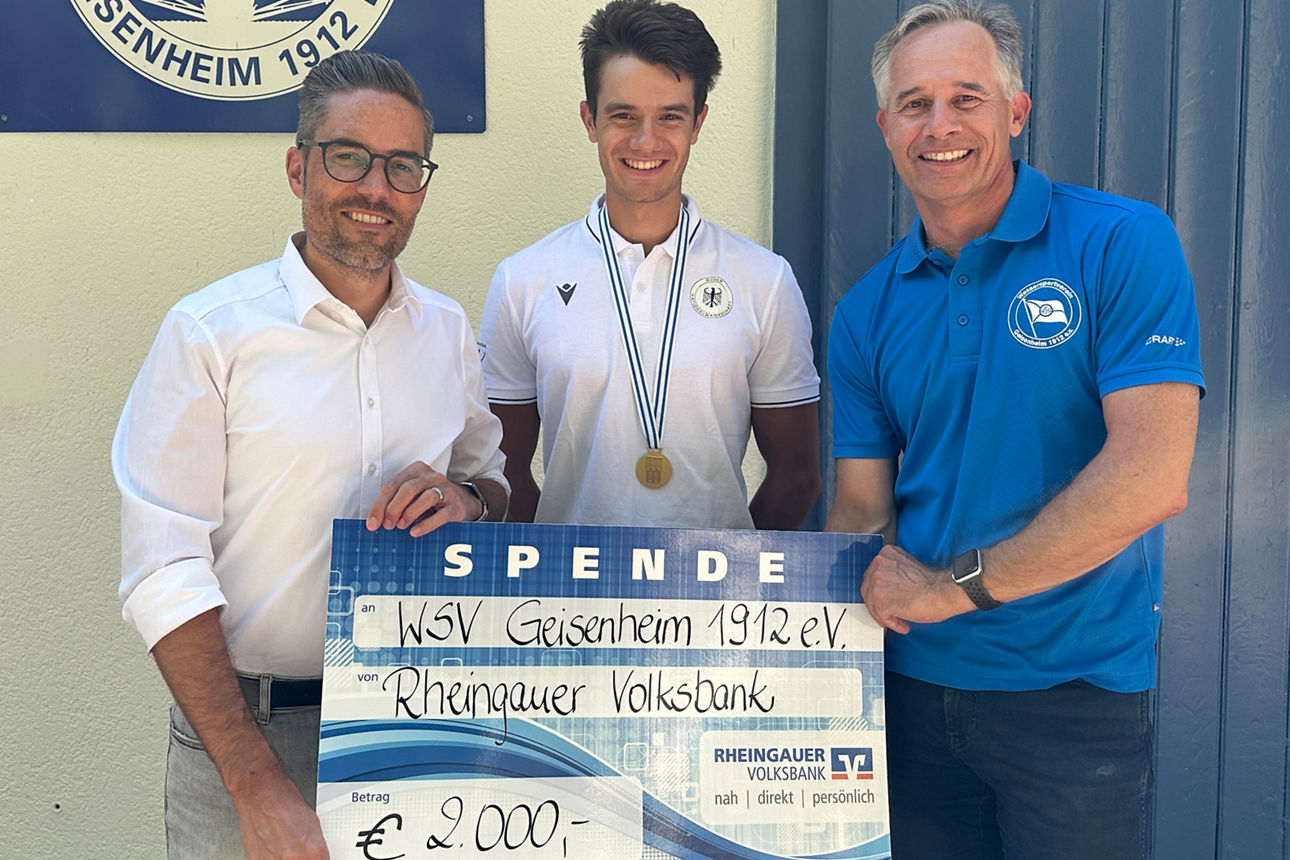 Drei Männer stehen vor einer Wand mit einem Schild des Wassersportvereins Geisenheim 1912 e.V., der mittlere trägt eine Medaille und hält einen großen symbolischen Scheck über 2000 Euro von der Rheingauer Volksbank.