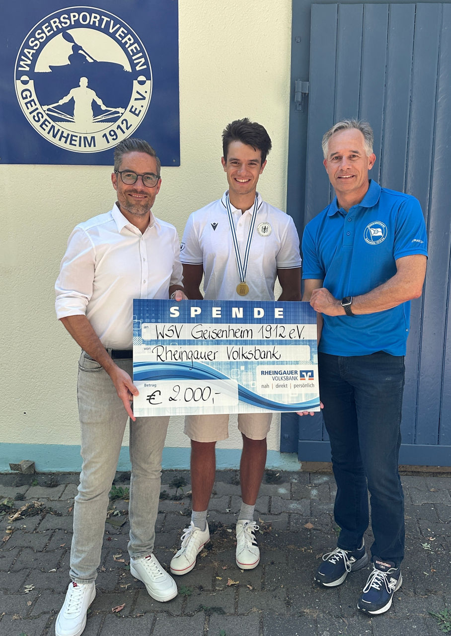 Drei Männer stehen vor einer Wand mit einem Schild des Wassersportvereins Geisenheim 1912 e.V., der mittlere trägt eine Medaille und hält einen großen symbolischen Scheck über 2000 Euro von der Rheingauer Volksbank.