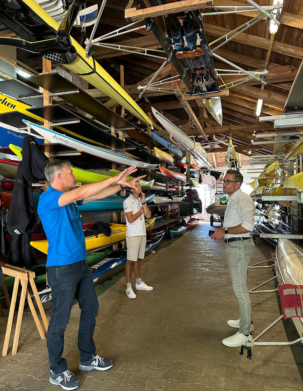 Drei Männer stehen in einem Bootshaus zwischen Regalen mit mehreren Kajaks und Kanus, einer erklärt etwas und zeigt mit ausgestreckten Armen