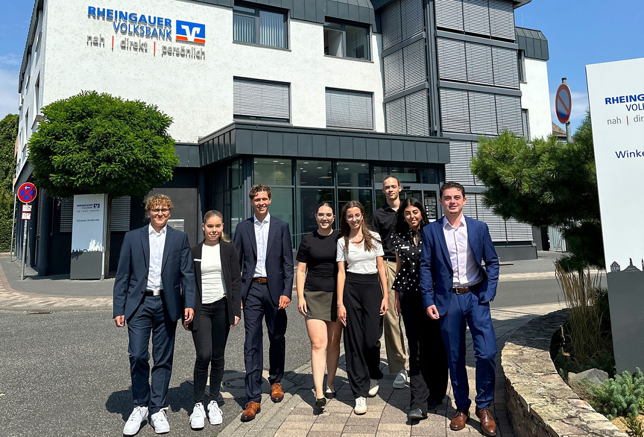 Neun junge Leute in Business-Kleidung gehen auf einem Bürgersteig. Im Hintergrund ein Bankgebäude