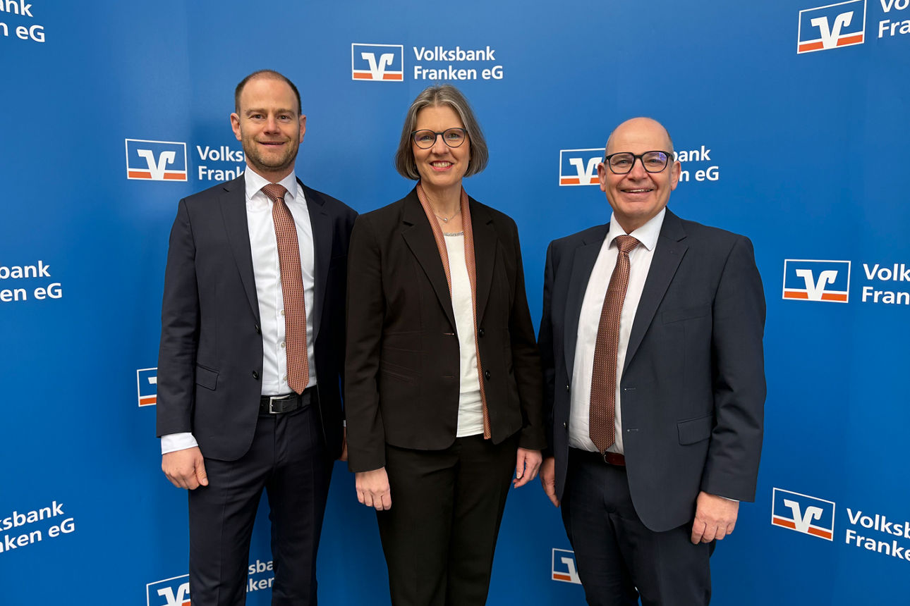 Die drei Vorstandsmitglieder stehen vor einer blauen Wand mit dem Logo der Volksbank Franken eG.