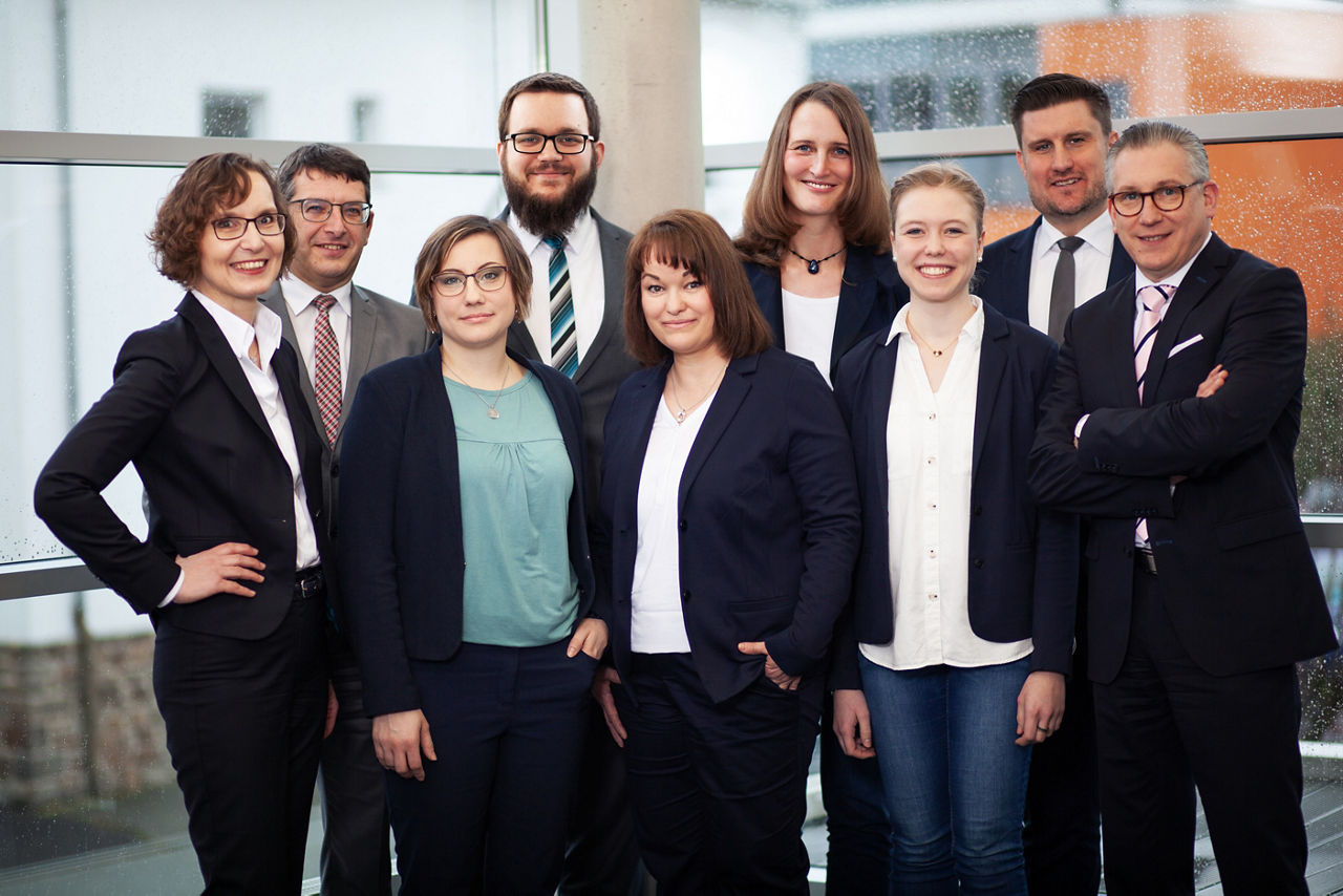 Das Team der SKB steht lächelnd vor einem Fenster
