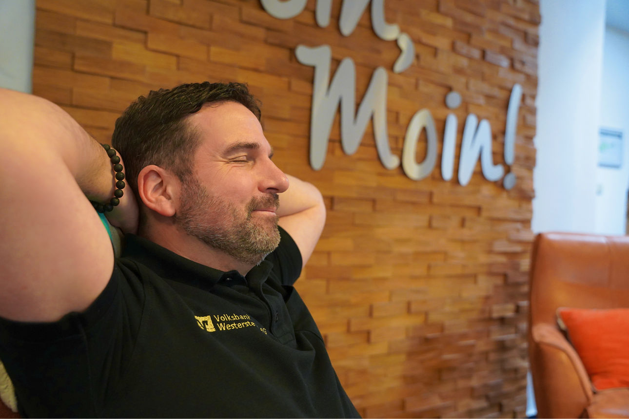 Ein Mitarbeiter der Volksbank Westerstede sitzt mit verschränkten Armen und Augen zu vor einem "Moin Moin"-Schild 