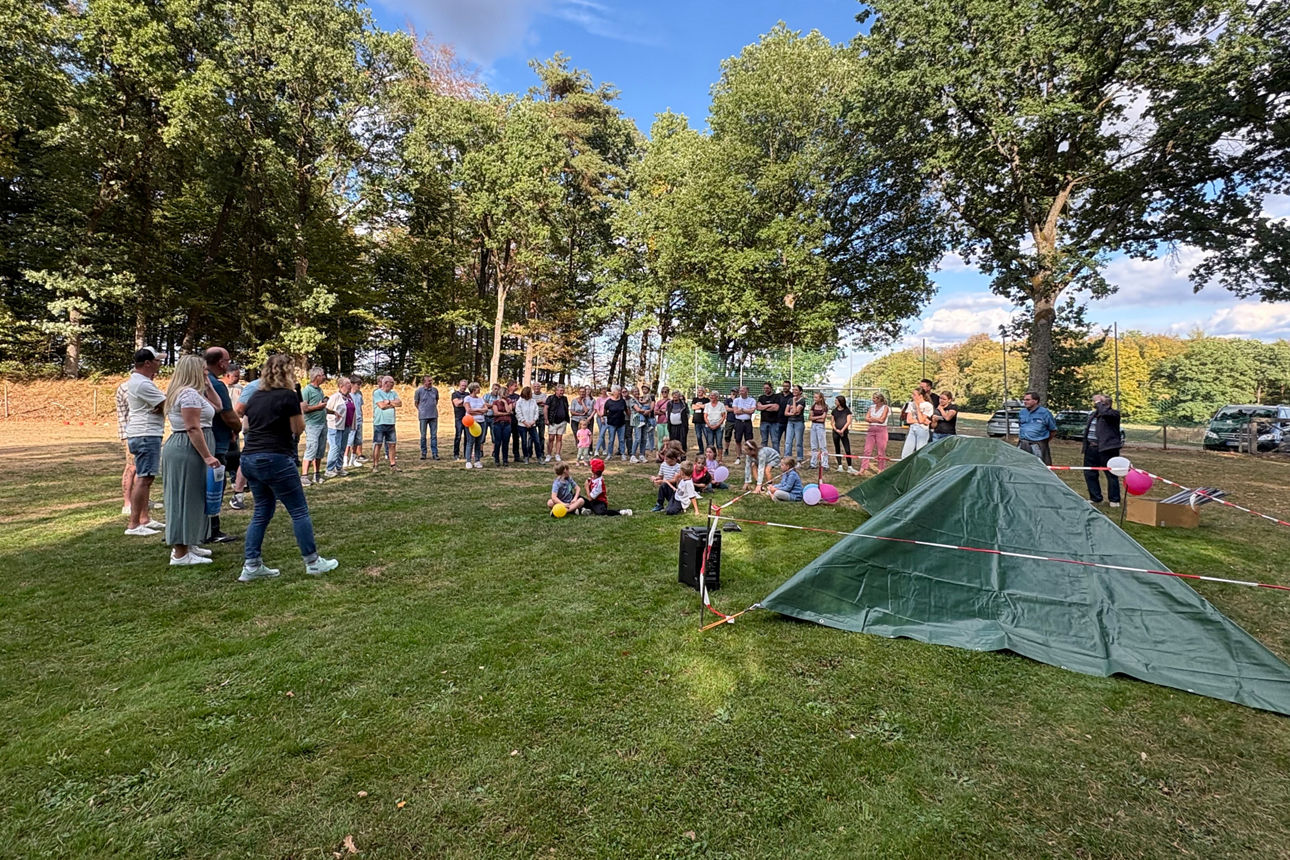 Menschen stehen auf einem Sportplatz um einen verhüllten Gegenstand, Kinder sitzen davor.