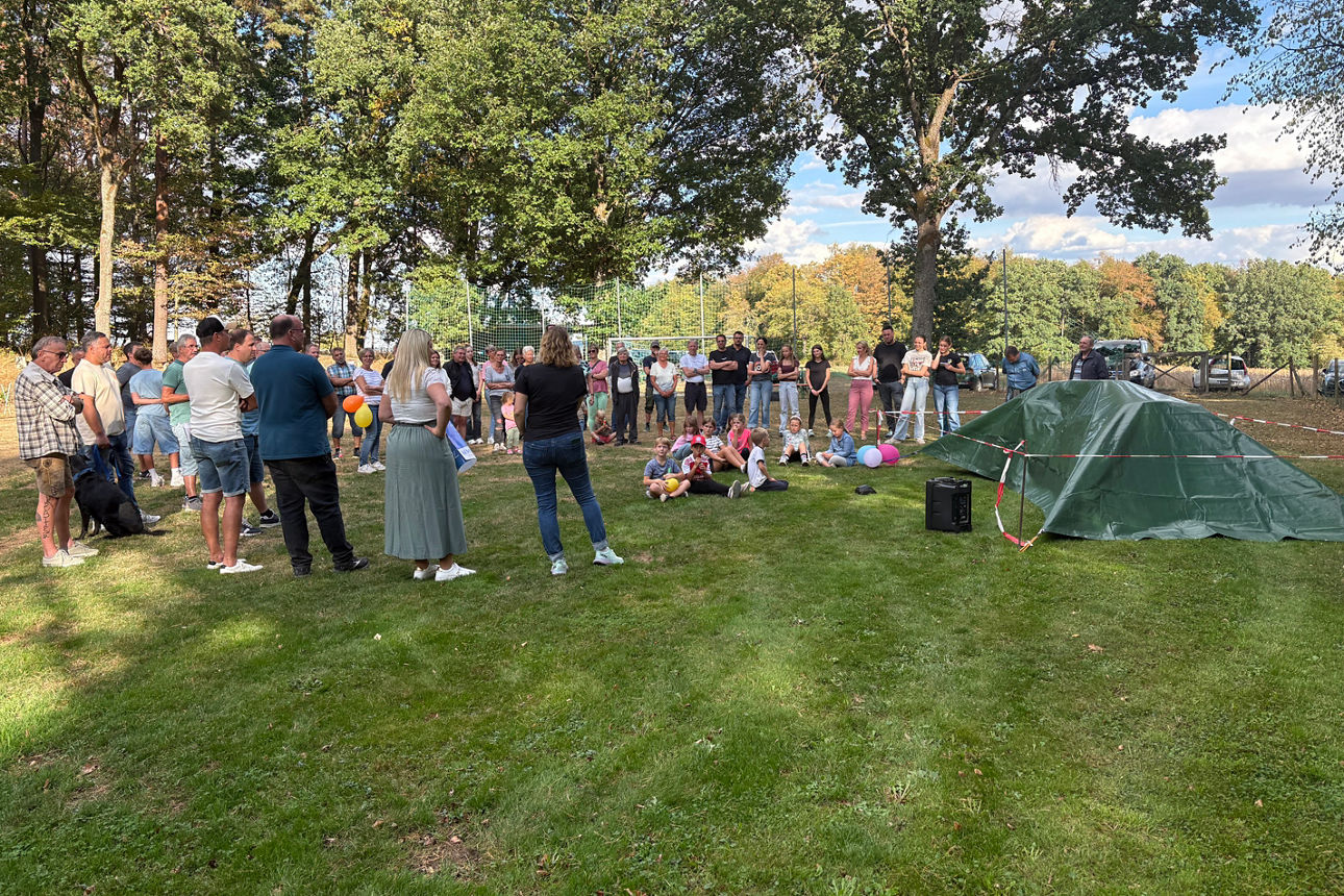 Menschen stehen auf einem Sportplatz um einen verhüllten Gegenstand, Kinder sitzen davor.