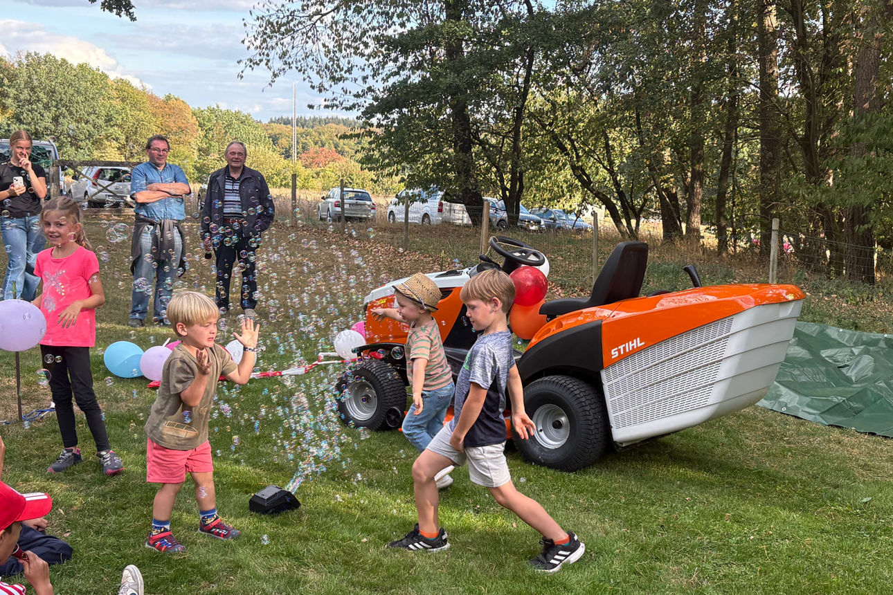 Kinder spielen mit Seifenblasen neben einem orangefarbenen Rasenmähertraktor auf dem Sportplatz.
