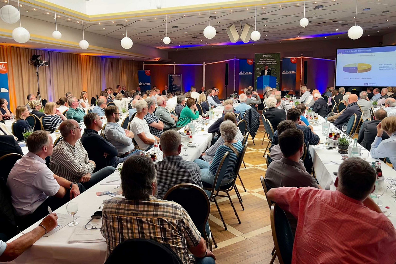 Ein großer Veranstaltungssaal voller Menschen, die an langen Tischen sitzen und einem Vortrag mit Leinwand-Präsentation folge