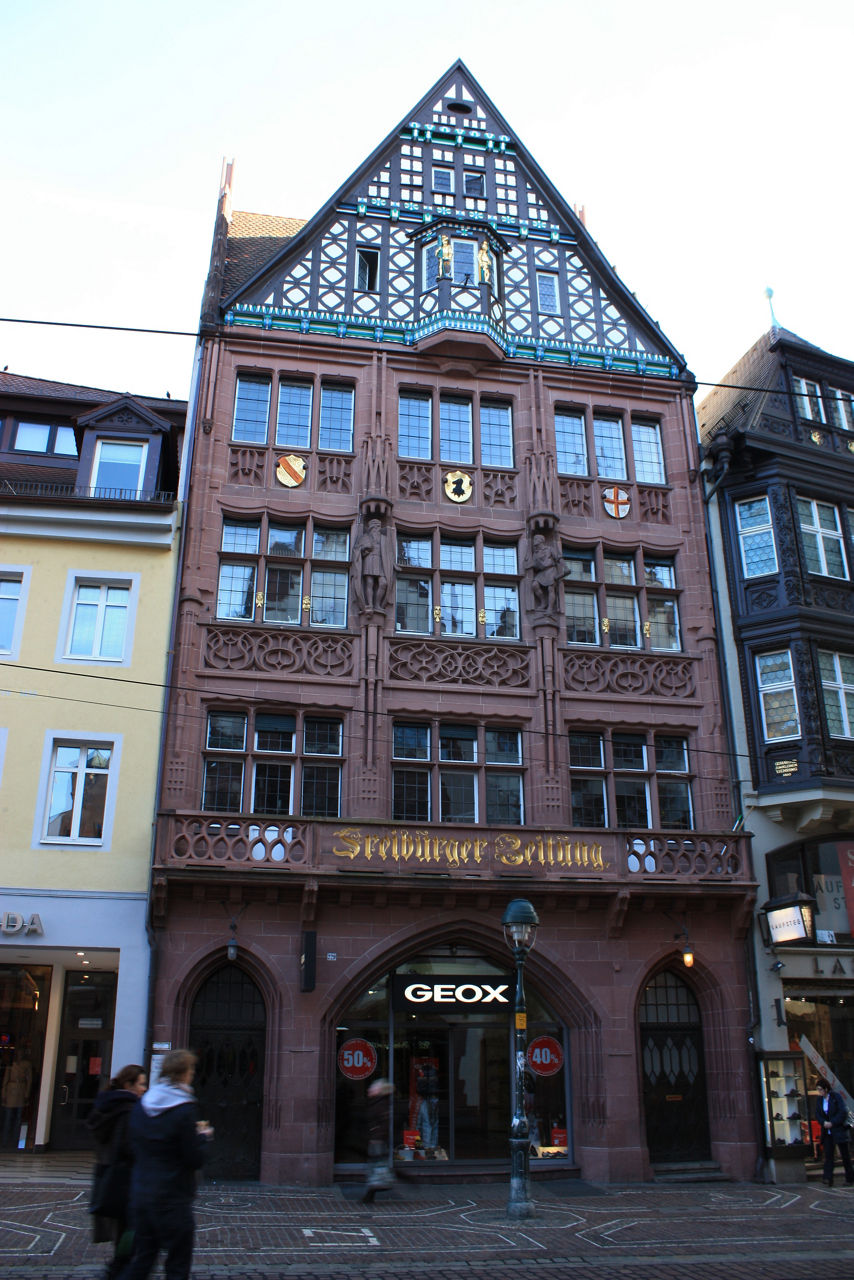 Auf dem Bild ist ein große, spitzes Gebäude mit vielen Fenstern zu sehen. Darauf steht Freiburger Zeitung in goldener Schrift.