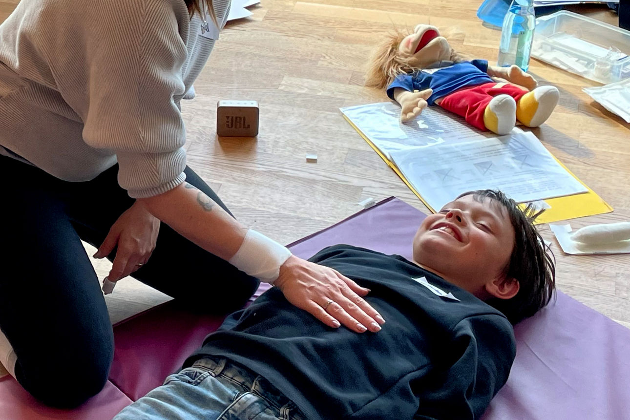 Kind liegt lächelnd auf einer Matte und lernt Erste-Hilfe-Maßnahmen
