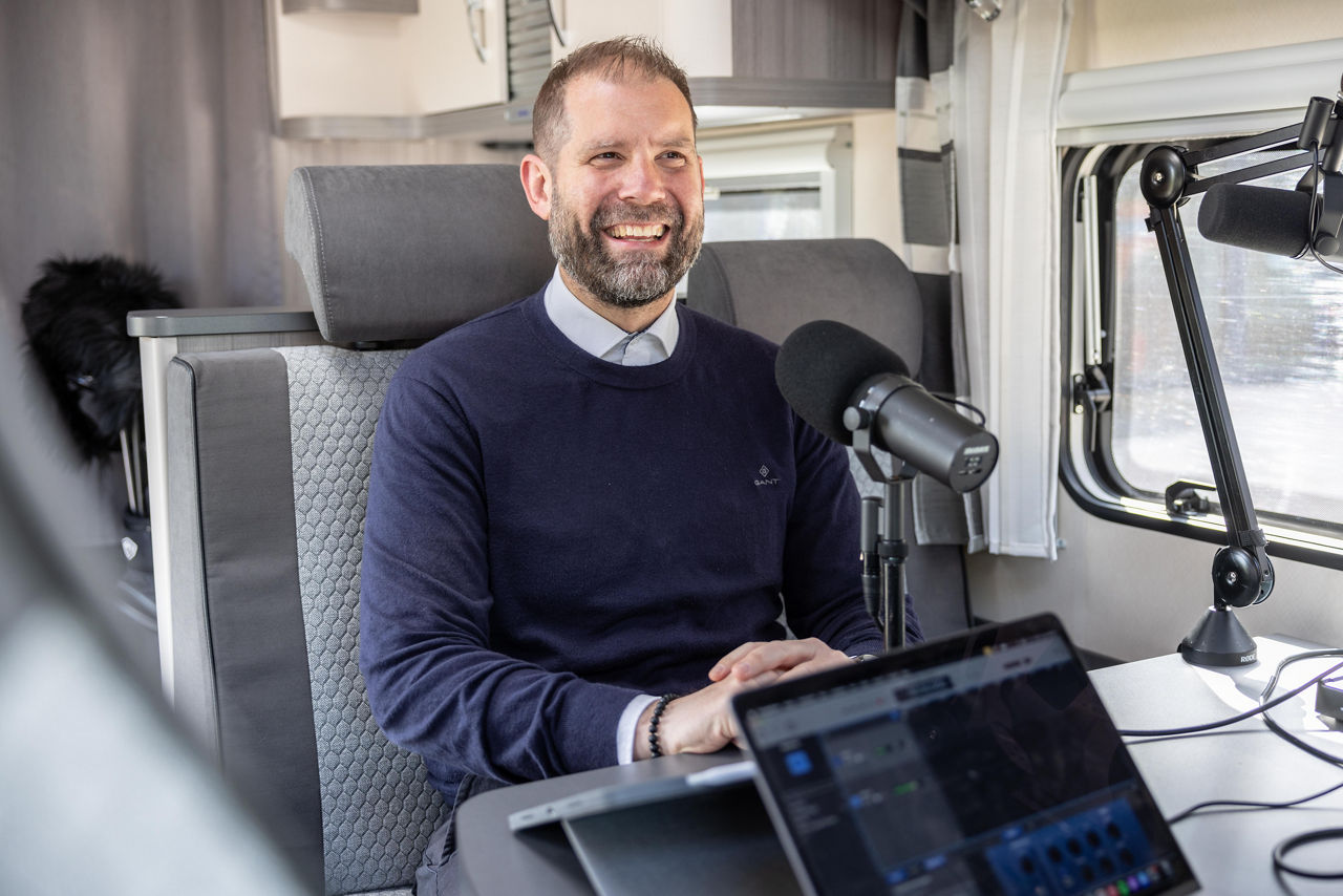 Ein Mann sitzt in einem Wohnmobil am Tisch und spricht in ein Mikrofon.