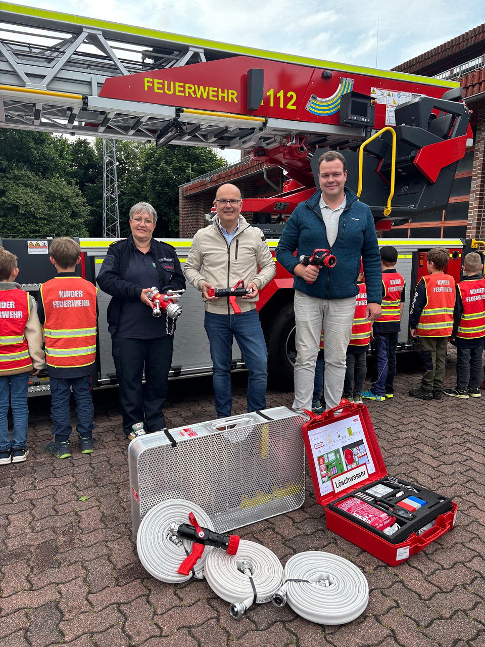 Drei Personen mit Sachspenden, im Hintergrund Kinder mit Warnwesten.