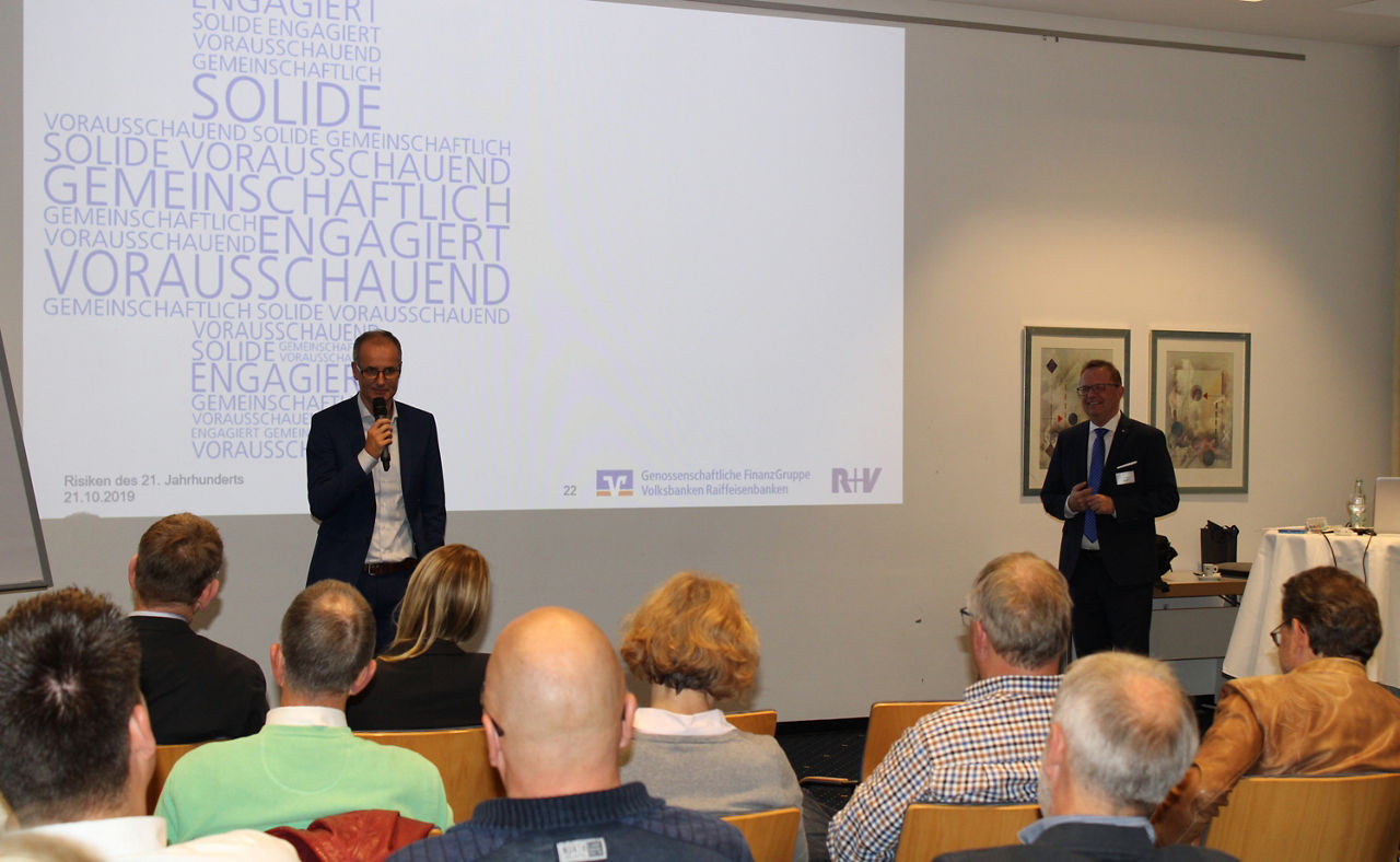 Die Präsentation findet in einem Konferenzraum statt, wobei ein Redner auf einem Podium sitzt und zu einem sitzenden Publikum