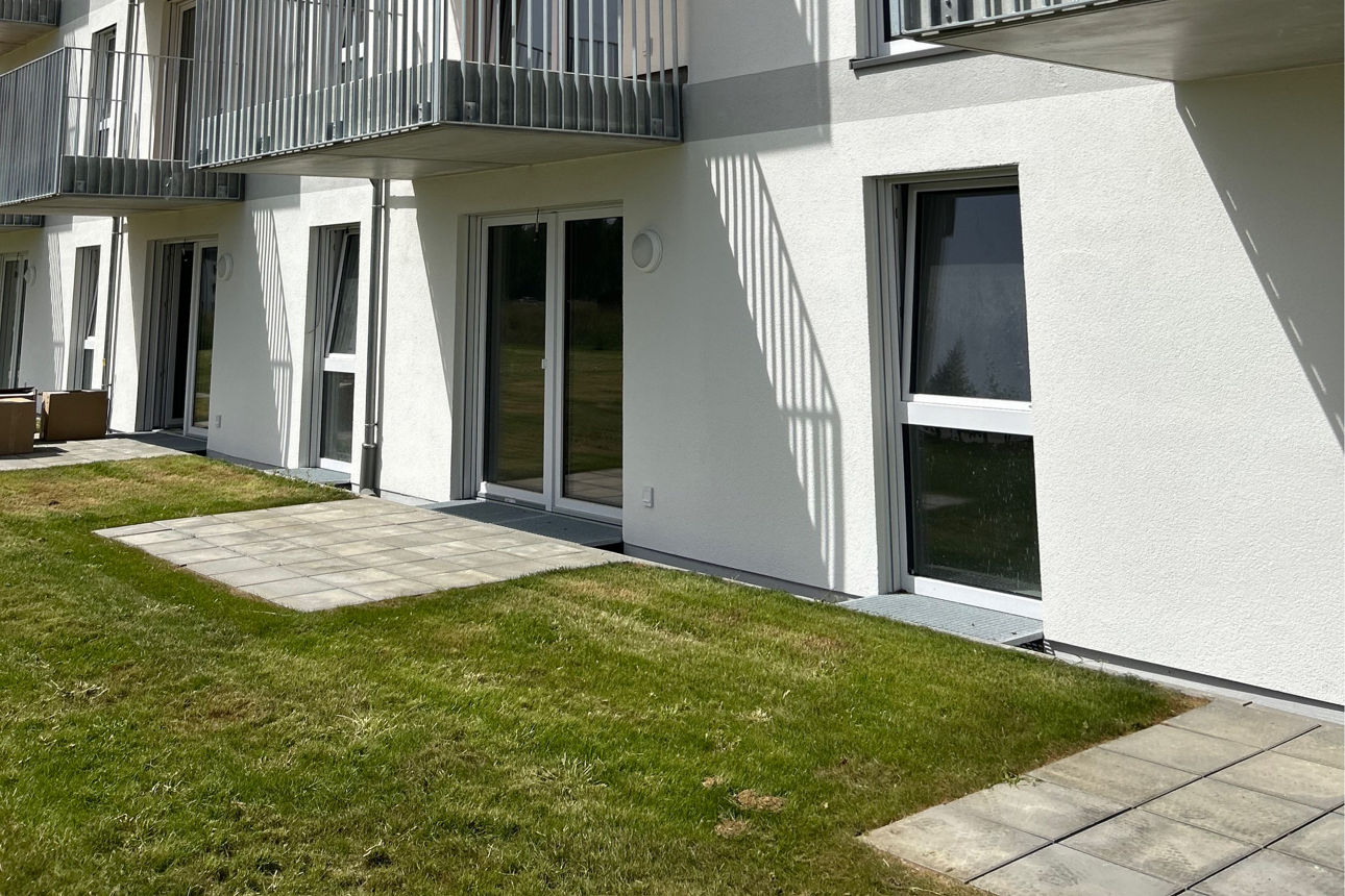 Weißes Neubau Mehrfamilienhaus. Davor eine grüne Wiese. Der Himmel ist blau. Es sind Balkone zu sehen.