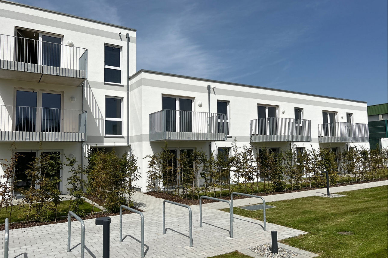 Weißes Neubau Mehrfamilienhaus. Davor eine grüne Wiese. Der Himmel ist blau. Es sind Fahrradstellplätze zu sehen.