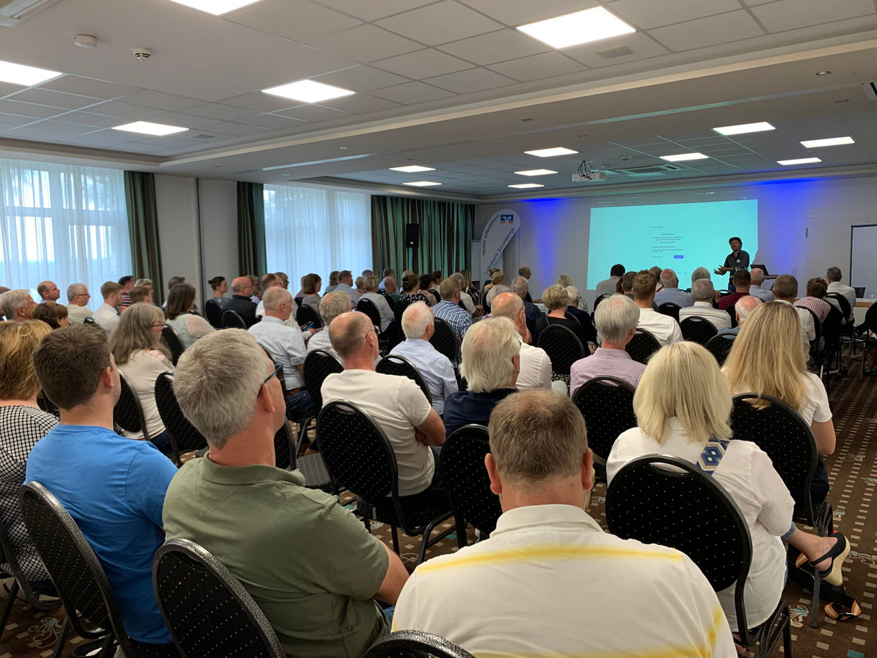 Eine große Gruppe von Personen sitzt in einem Konferenzraum und hört sich eine Präsentation an.