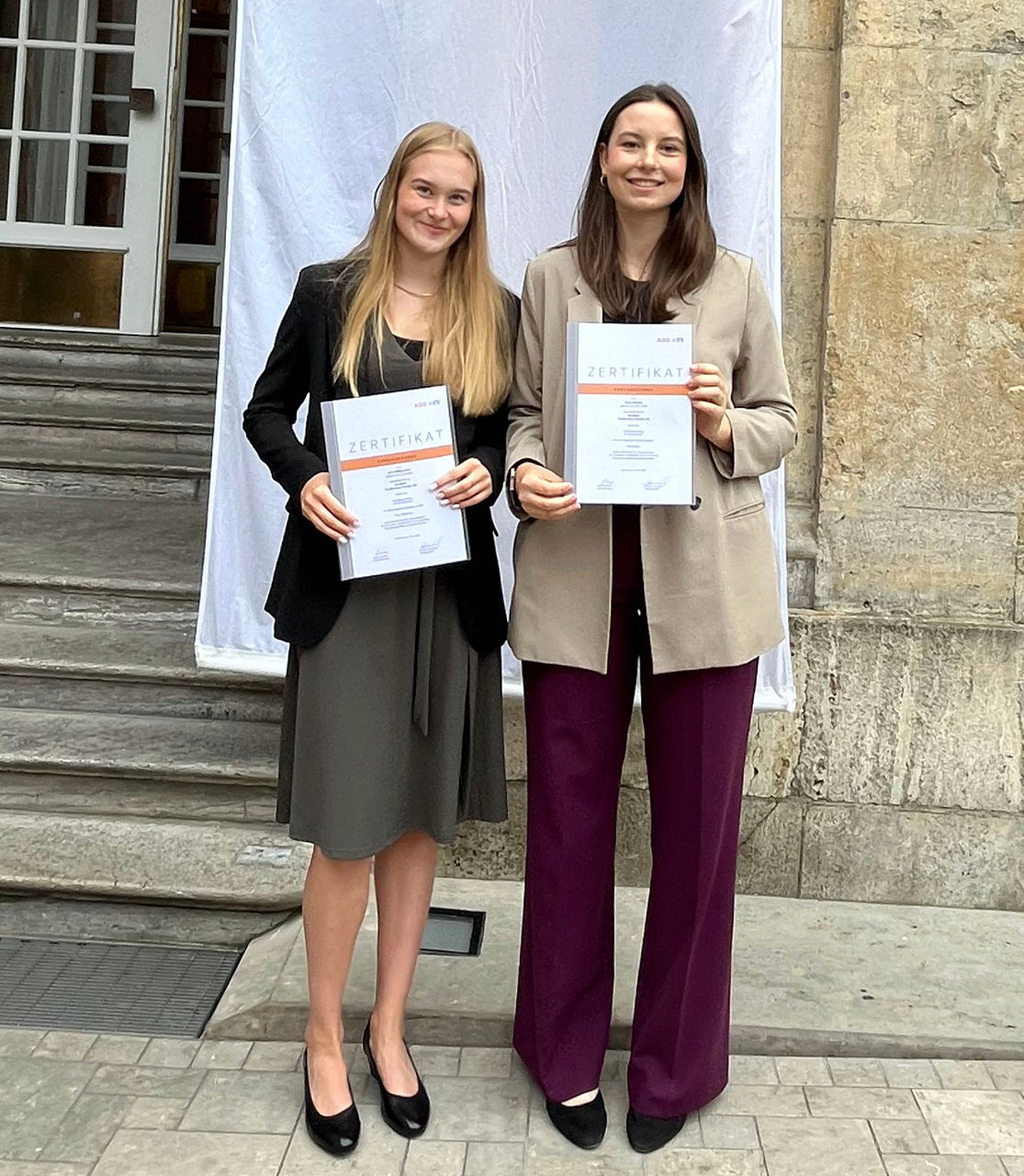 Zwei junge Frauen stehen mit Zeugnissen in der Hand vor einem schulischen Gebäude nebeneinander und lächeln in die Kamera. 