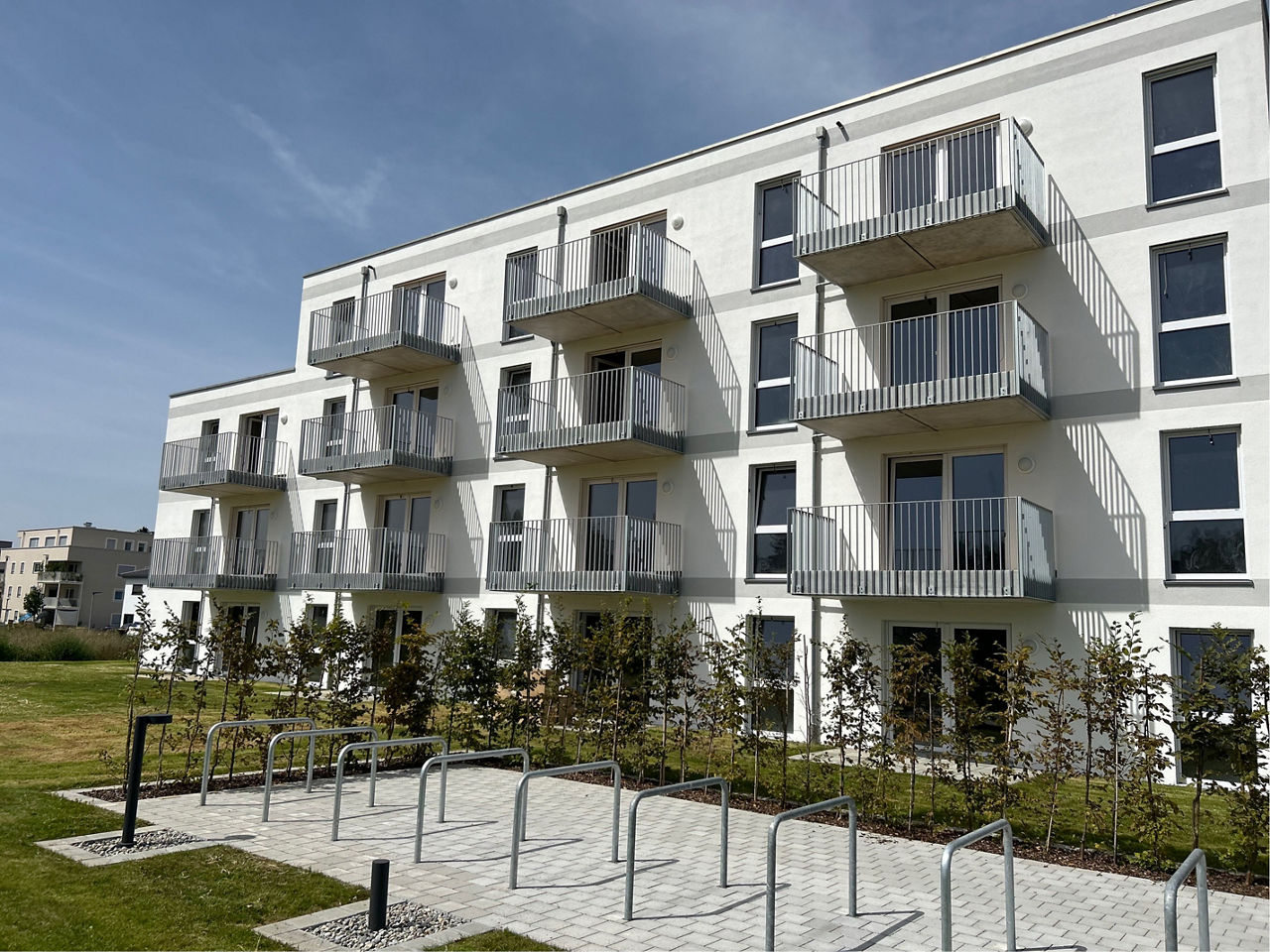 Weißes Neubau Mehrfamilienhaus. Davor eine grüne Wiese. Der Himmel ist blau. Es sind Fahrradstellplätze zu sehen.