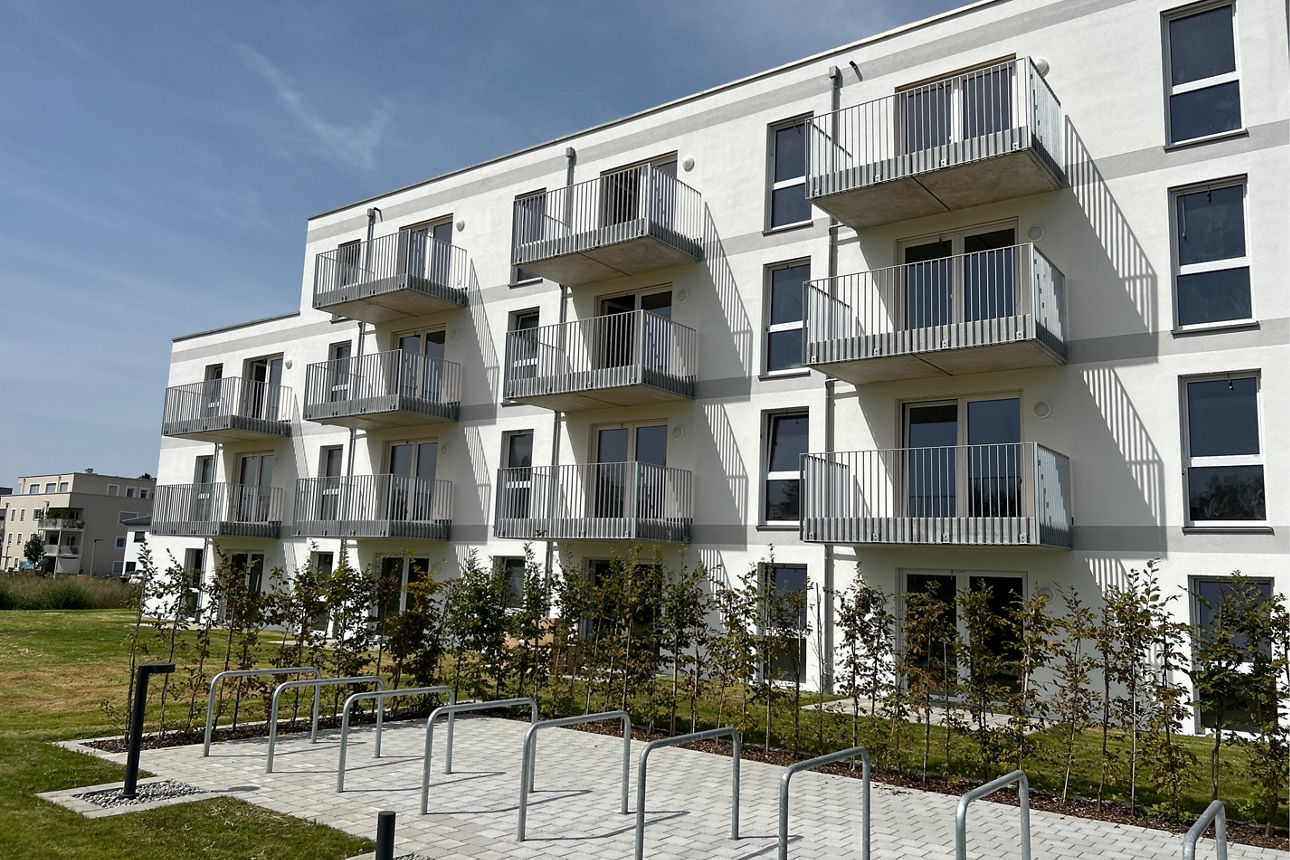 Weißes Neubau Mehrfamilienhaus. Davor eine grüne Wiese. Der Himmel ist blau. Es sind Fahrradstellplätze zu sehen.