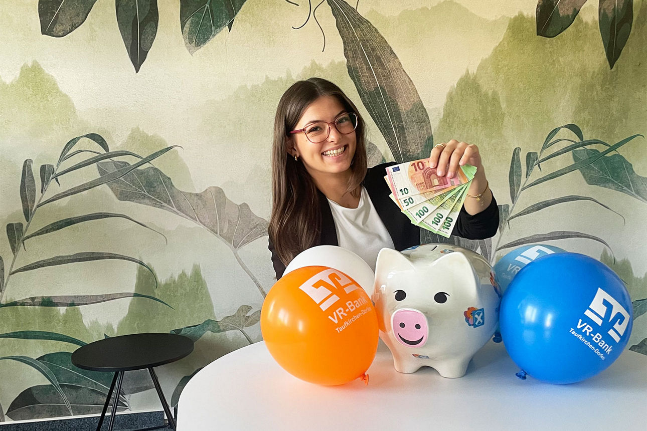 junge Frau sitzt mit 360 € in der Hand vor einer bunten Tapete und lächelt. Vor ihr sind Luftballons und ein Sparschwein
