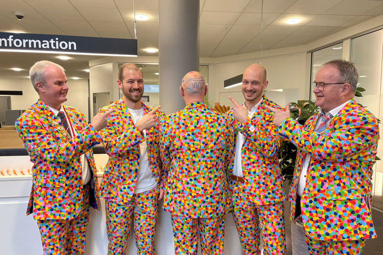 Fünf Mitarbeiter der Raiffeisenbank tragen Konfetti-Kostüme, vier zeigen auf einen Geschminkten Hinterkopf mit Raiba-Logo