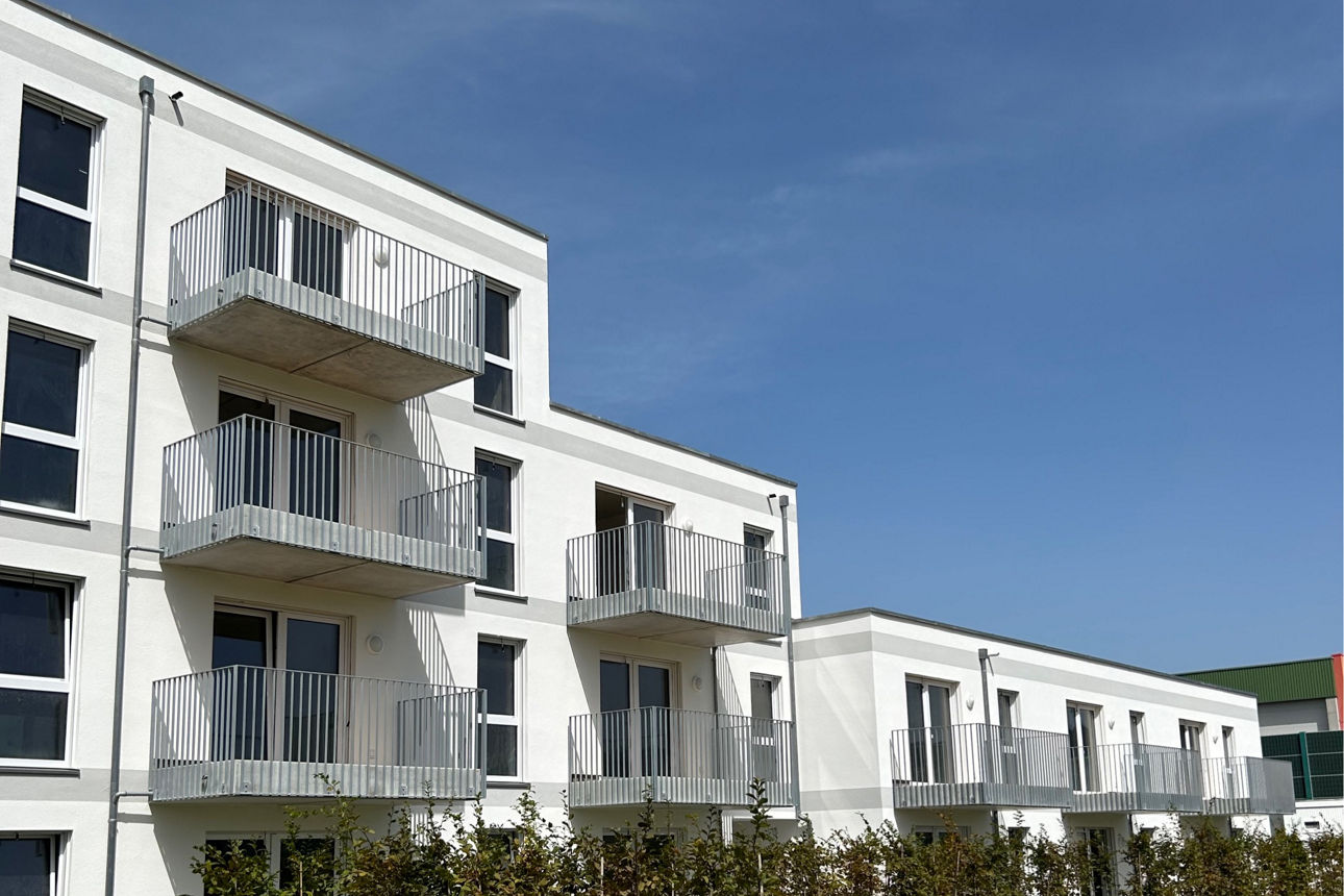 Weißes Neubau Mehrfamilienhaus. Davor eine grüne Wiese. Der Himmel ist blau. Es sind Fahrradstellplätze zu sehen.