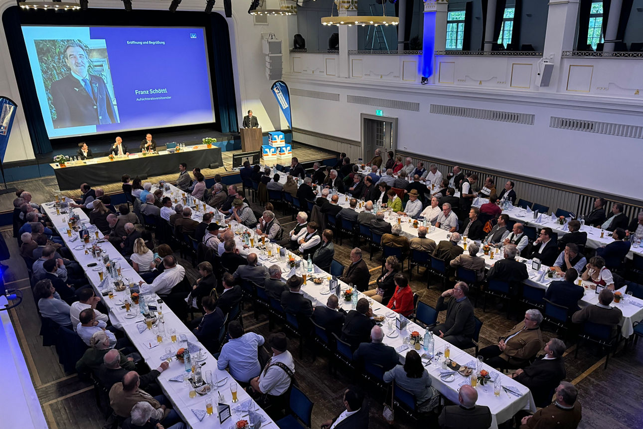 Gesamtansicht einer großen Veranstaltungshalle mit vielen Gästen, die an langen Tischen sitzen. Eine Bühne mit Rednerpult und großer Leinwand im Hintergrund zeigt das Porträt eines Mannes. Die Decke ist mit Beleuchtung und großen Kronleuchtern versehen.