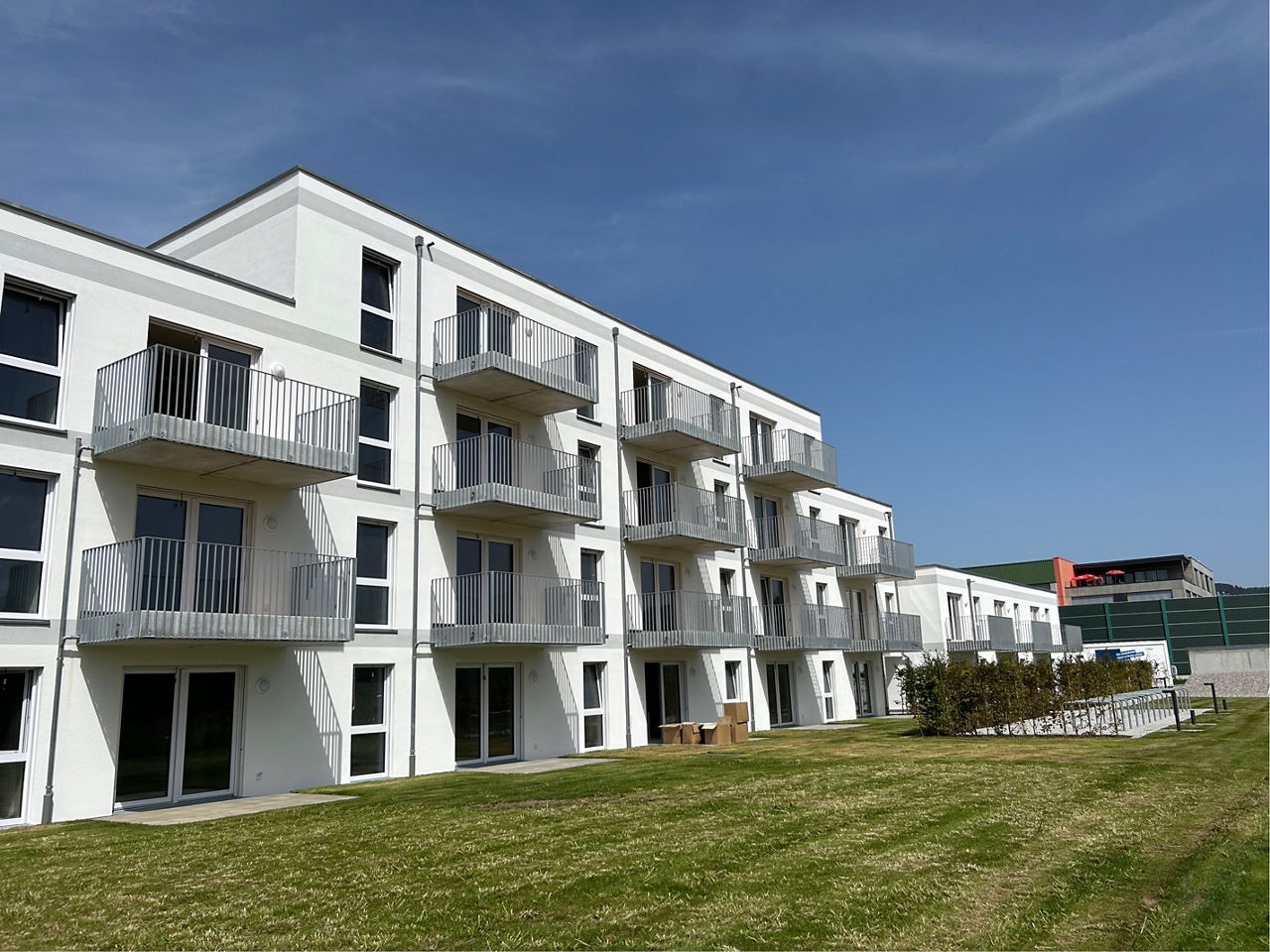 Weißes Neubau Mehrfamilienhaus. Davor eine grüne Wiese. Der Himmel ist blau. Es sind Balkone zu sehen.