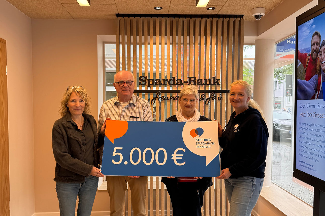 Vier Personen stehen in einem Raum und halten einen Spendenscheck über 5.000 Euro