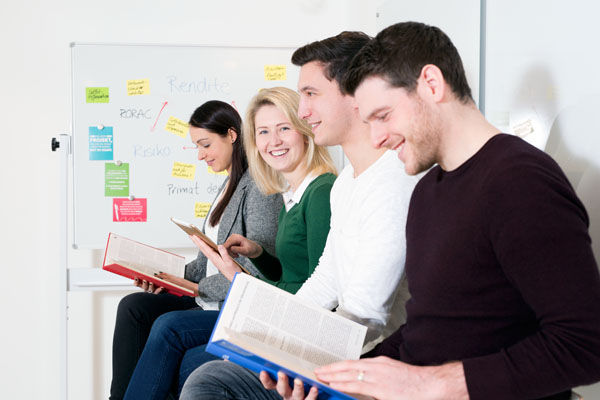 Vier junge Personen sitzen in einem Besprechungsraum an der Wand gelehnt und schauen sich Bücher an.