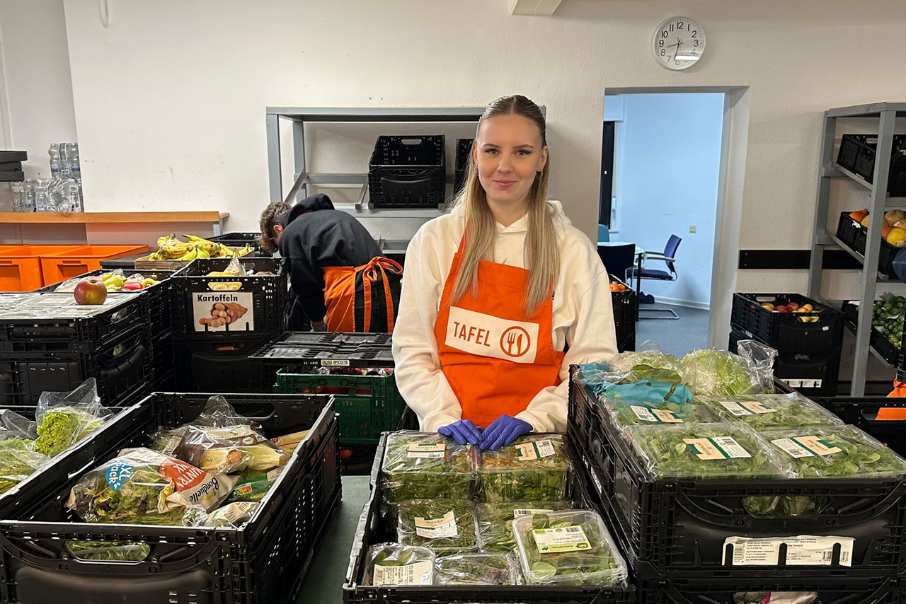 Eine junge Tafel-Helferin steht mit orangefarbener Schürze zwischen Kisten mit Salat, Gemüse und weiteren Lebensmitteln.