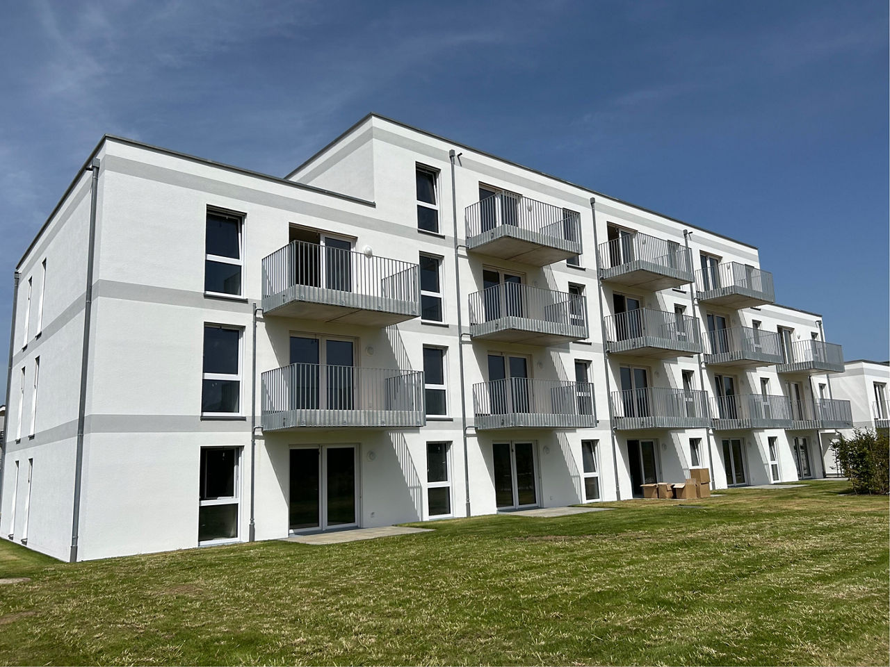 Weißes Neubau Mehrfamilienhaus. Davor eine grüne Wiese. Der Himmel ist blau.