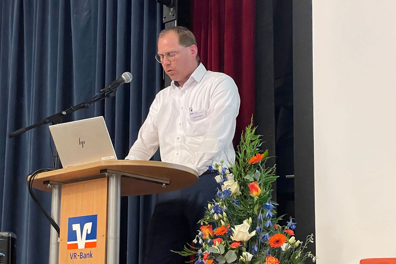 Mann in weißem Hemd steht auf einer Bühne vor Rednerpult und spricht zum Publikum.