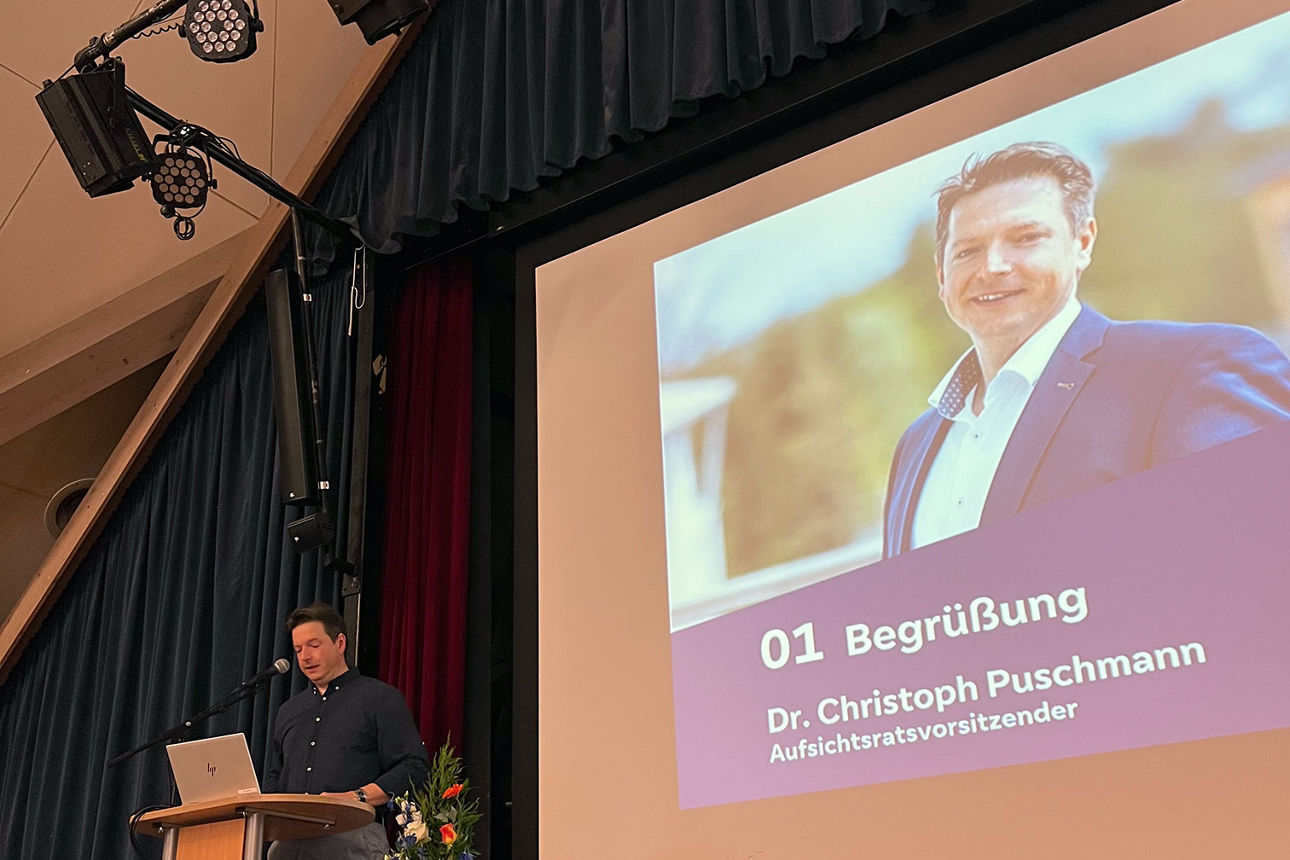 Mann hält Begrüßungsrede auf einer Bühne mit Rednerpult und großem Beamerbildschirm