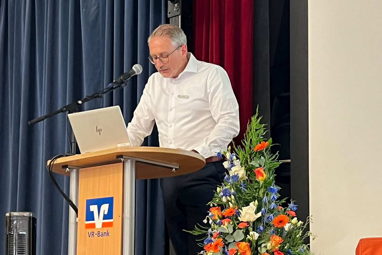 Mann steht neben Blumenschmuck auf Bühne vor Rednerpult und spricht zum Publikum.