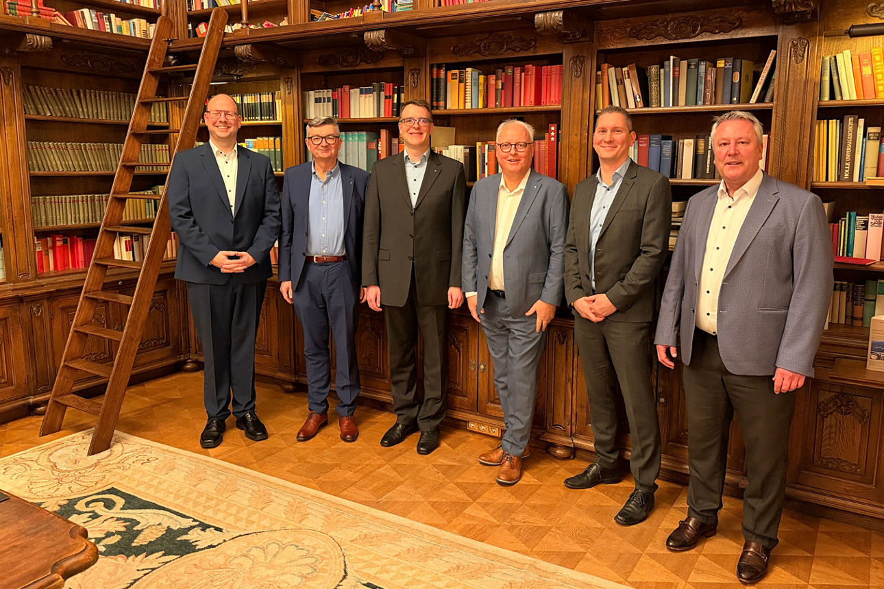 Sechs Männer stehen in einer Bibliothek vor gefüllten Bücherregalen und posieren gemeinsam für ein Gruppenfoto.