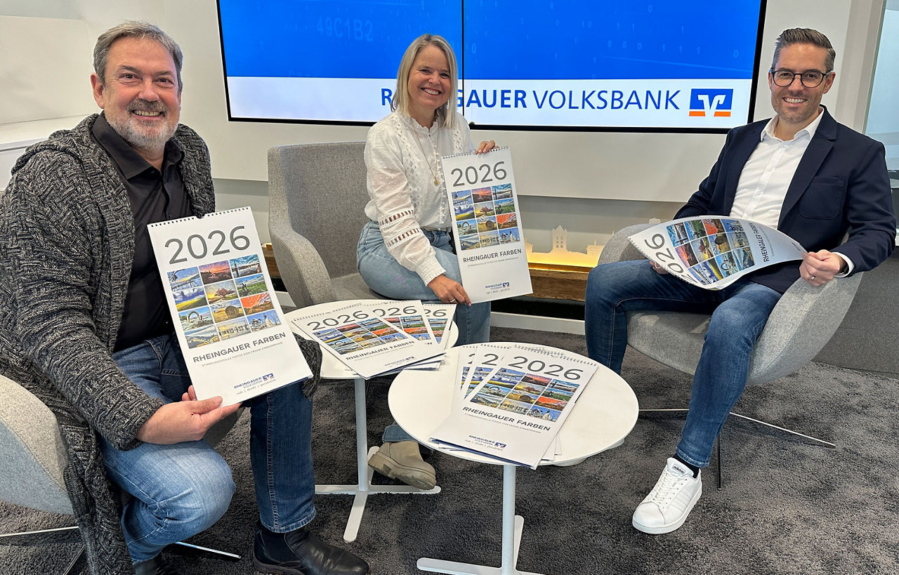 Drei Personen sitzen in einem modernen Raum, halten Kalender mit dem Titel '2026' und dem Schriftzug 'Rheingauer Volksbank' auf einem Bildschirm im Hintergrund.