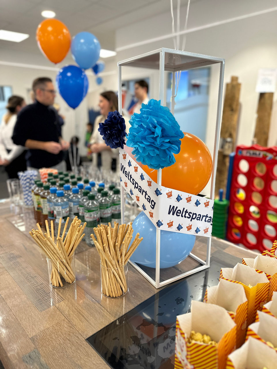 Dekorierter Tisch mit blauen und orangen Luftballons, Papierblumen, Popcorn, Getränken und einem Schild mit der Aufschrift 'Weltspartag'.