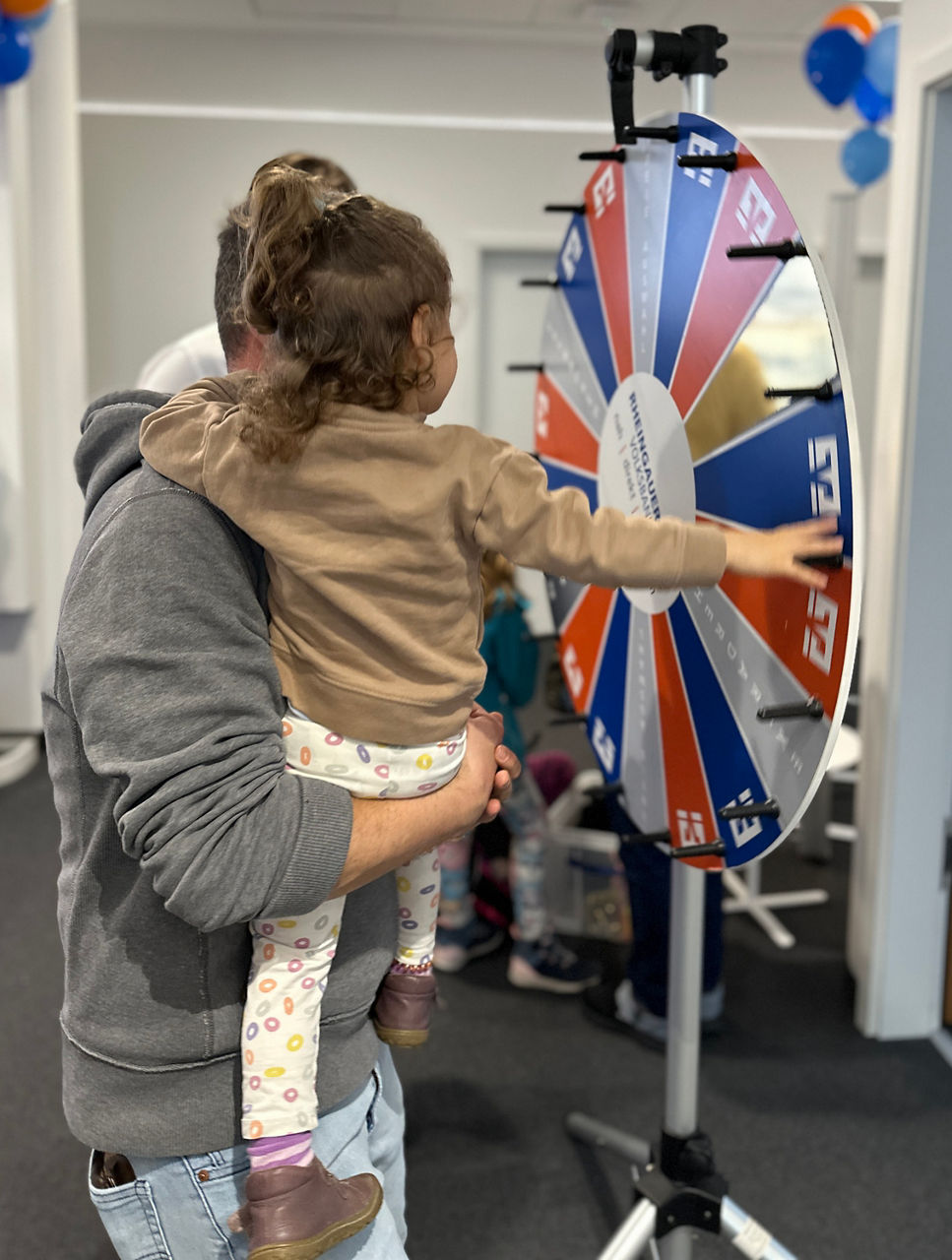 Ein Erwachsener hält ein Kleinkind, das ein buntes Glücksrad mit roten, blauen und weißen Segmenten dreht.