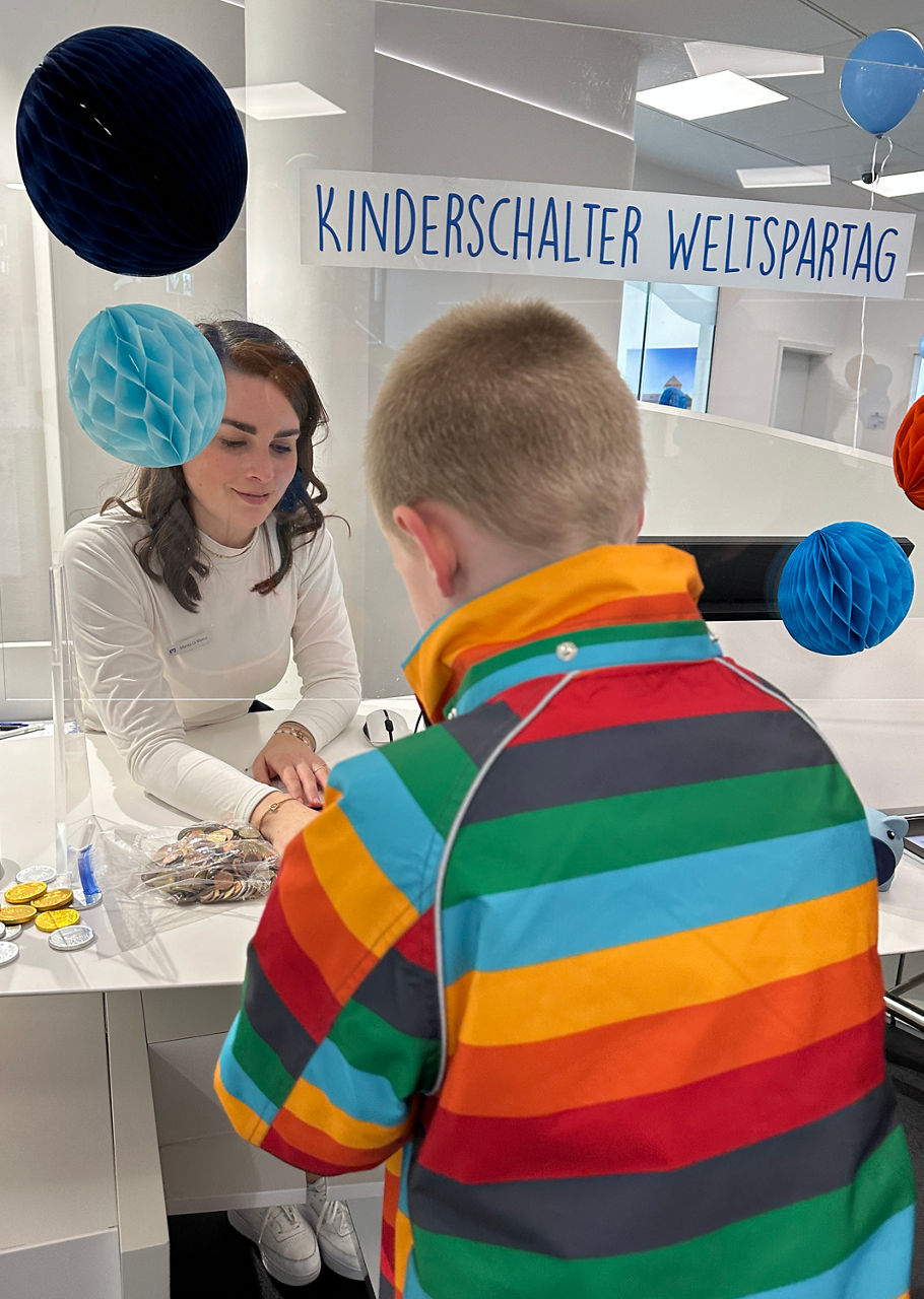Kind mit bunter Regenbogenjacke steht an einem weißen Tisch, an dem eine Frau sitzt, im Hintergrund ein Schild mit der Aufschrift 'KINDERSCHALTER WELTSPARTAG' und bunte Papierdekorationen.