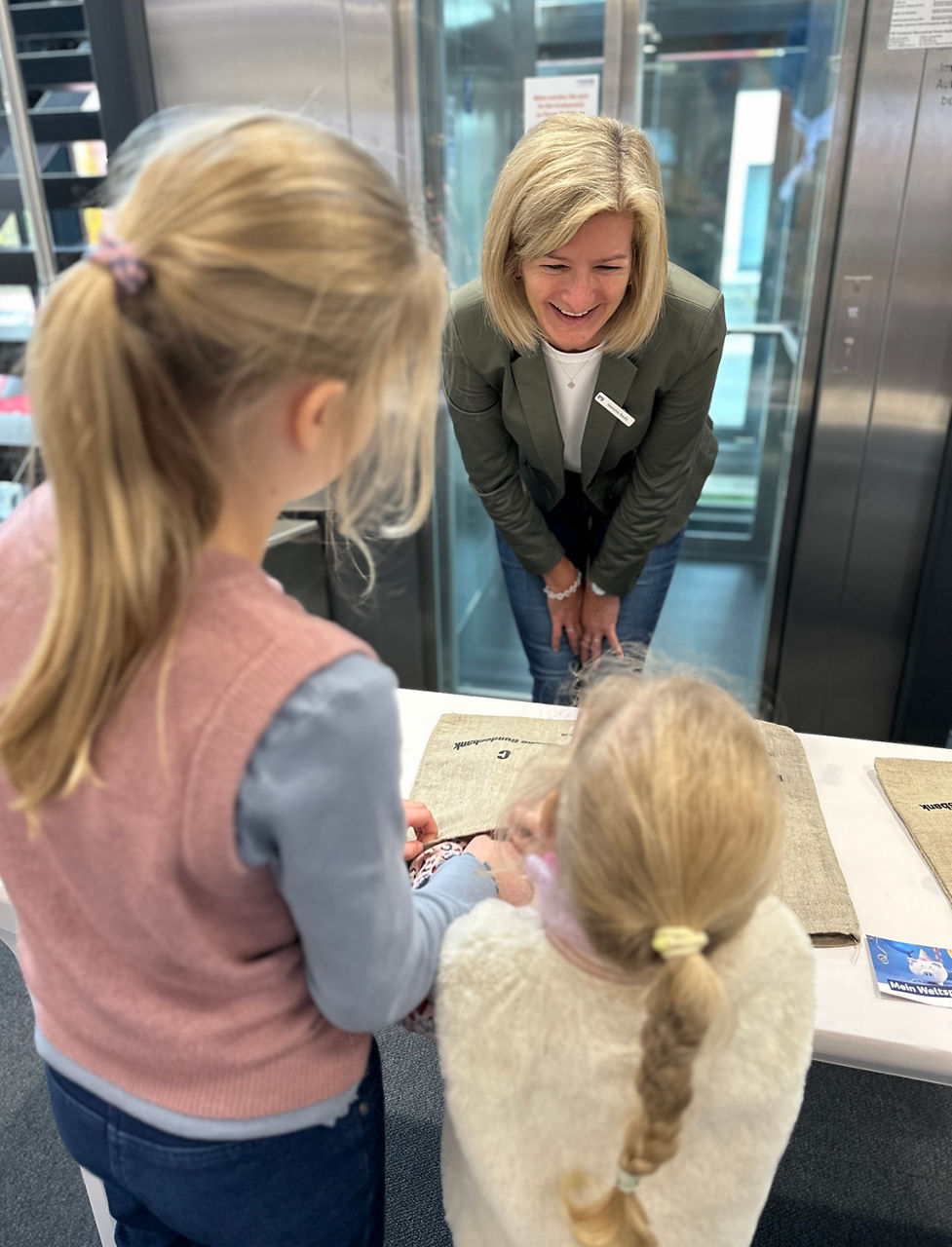 Frau mit Namensschild beugt sich über Tisch, zwei Mädchen mit Zöpfen stehen davor, im Hintergrund Aufzugtüren.