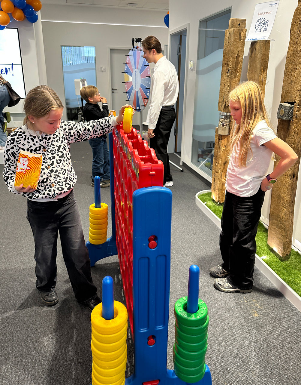 Zwei Kinder spielen an einem großen, roten Vier-gewinnt-Spiel mit gelben und grünen Spielsteinen in einem Innenraum.