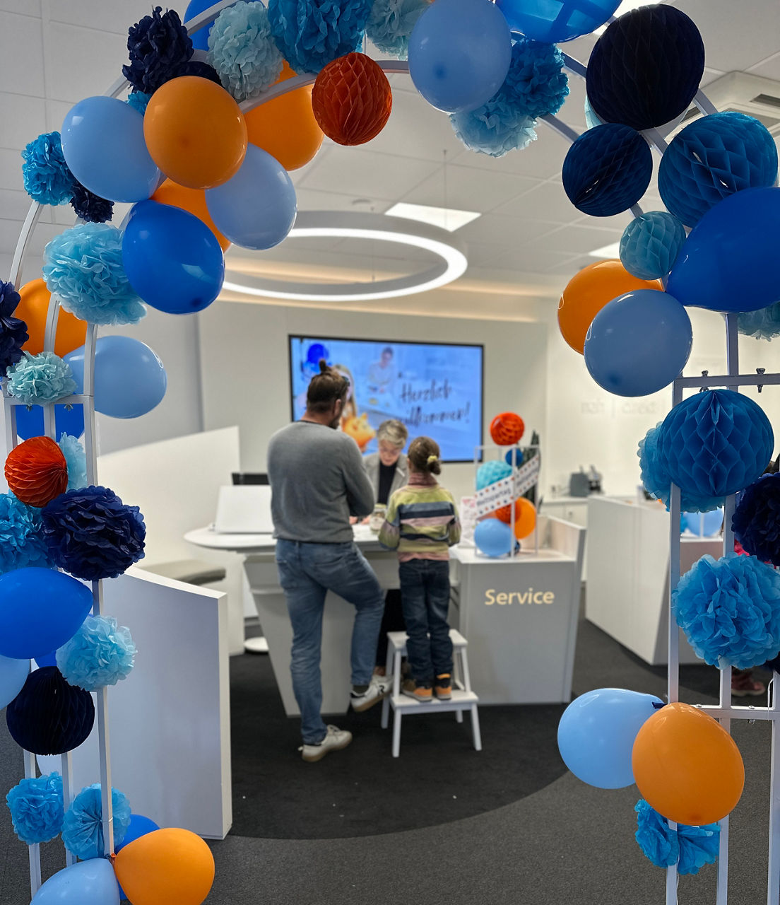 Bunte Luftballons und Papierblumen in Blau- und Orangetönen rahmen einen Mann und ein Kind, die vor einem Bildschirm in einem modernen Servicebereich stehen.